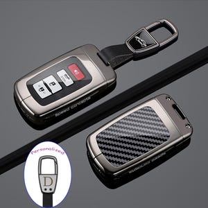 May include: A gunmetal gray car key fob cover with a carbon fiber pattern on the back and a matching metal keychain attachment. The fob features the text Speed Concepts on the side. An inset shows a personalized keychain with the letter D.