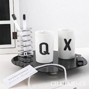 Può includere: Un moderno set da bagno con due tazze in ceramica bianca con disegni di lettere nere, un portaspazzolino argentato con due spazzolini e un vassoio nero. Sono presenti anche una collana di perle e un'etichetta con testo.