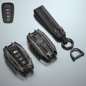 May include: A black car key fob cover with a mechanical design, featuring a matching black leather strap embossed with the word PERSONALIZED, a circular metal ring, and a D-shaped shackle. The cover has cutouts for buttons and a shield logo.