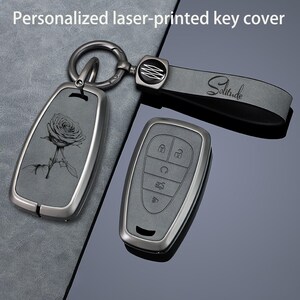Può includere: Un coprichiave grigio scuro con un disegno di rosa inciso al laser, fissato a un anello di metallo e a un cinturino grigio con la parola Solitude. Un telecomando per auto grigio scuro abbinato con cinque pulsanti è posto accanto.