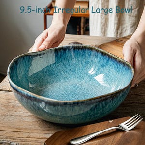 May include: A 9.5-inch irregular large bowl with a reactive glaze finish in shades of blue and teal, featuring a dark rim. The bowl has a speckled texture and a wide, open basin. A silver-toned metal fork is placed on a wooden surface nearby.
