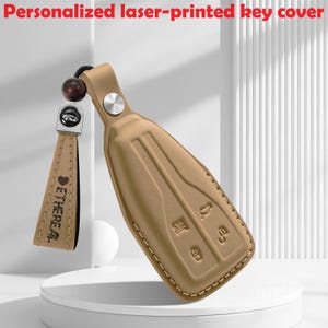 Puede incluir: Una funda de cuero color canela para llave de coche con iconos de botones en relieve y una correa a juego con el texto grabado con láser "ETHEREAL" y un símbolo de corazón. La funda se asegura con un remache de metal plateado y una cuenta de madera.