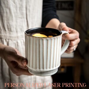 May include: A white ceramic mug with vertical ribbed detailing and a black interior, held by a person. The mug has a rounded base and a curved handle. The mug is filled with a beverage and a slice of lemon. Text at the bottom reads "PERSONALIZED LASER PRINTING."