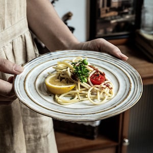 Peut inclure: Une assiette ronde en céramique à la surface crème, présentant des stries circulaires concentriques et un bord brun foncé. L'assiette contient une portion de spaghettis garnie d'un quartier de citron, d'une tomate cerise, de petits pois et de persil.