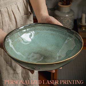 Può includere: Una grande ciotola in ceramica color verde acqua con smalto maculato e bordo marrone scuro. La ciotola è tenuta da una persona che indossa un grembiule beige. Il testo "PERSONALIZED LASER PRINTING" è visibile in basso.