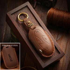 Pode incluir: Uma capa de couro castanho para chave de carro com costuras visíveis, apresentando um anel de metal cor de latão e uma alça de couro. A inserção mostra a mesma capa com a palavra UNIQUE gravada sob o texto PERSONALIZATION.