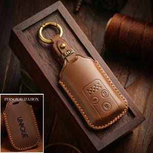 May include: A brown leather key fob case with a gold-colored ring and clasp. The case has embossed buttons and stitching. A smaller image shows a similar case with the word "UNIQUE".