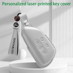 May include: A light grey car key cover featuring embossed button icons and a matching leather strap with the word PERSONALIZED printed on it. The strap is attached to the cover with a silver metal stud and a dark brown wooden bead.