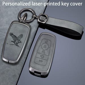May include: A grey personalised laser-printed key cover with an eagle graphic, featuring a matching grey strap and a dark metal ring. The key cover has a rectangular shape with rounded corners and includes a set of buttons for vehicle control.