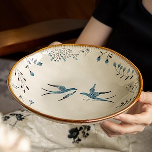 Puede incluir: Un cuenco de cerámica redondo con base color crema y borde marrón oscuro. El interior presenta un diseño pintado a mano de dos golondrinas azules volando una hacia la otra, rodeadas de delicadas ramitas florales y ramas con hojas.