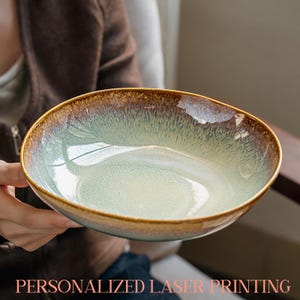 Op de afbeelding: Een keramische kom met een unieke glazuur. De kom heeft een lichtgroen interieur, dat overgaat in een bruine rand. De tekst "PERSONALIZED LASER PRINTING" staat onderaan de afbeelding.