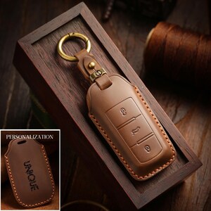 May include: A brown leather car key fob cover with a gold-colored keyring. The cover has three button icons and is displayed in a wooden box. The word "UNIQUE" is embossed on a similar cover in the corner.