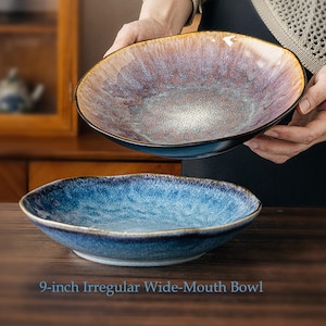 Puede incluir: Dos cuencos de boca ancha e irregular de 23 cm con acabado de esmalte reactivo. Un cuenco presenta un degradado en tonos marrón, cobre y crema, mientras que el otro muestra un patrón moteado en azul oscuro y claro con bordes oscuros.