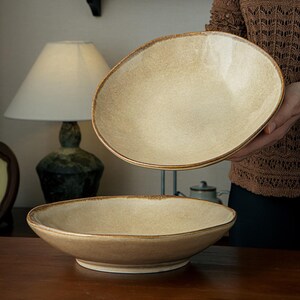 May include: Two shallow, round ceramic bowls with a speckled, light beige glaze and a darker, uneven rim. The bowls feature a smooth, organic shape with a slightly irregular edge, showcasing a natural, earthy finish on the interior and exterior.