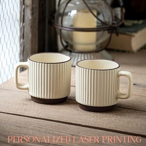 May include: Two cream-colored ceramic mugs with vertical ribbed detailing and dark brown bases and rims. Each mug has a handle and sits on a wooden surface. The text "PERSONALIZED LASER PRINTING" is at the bottom of the image.