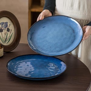 Peut inclure: Deux assiettes rondes en céramique dotées d'un émail réactif bleu profond avec une texture mouchetée subtile et un bord sombre irrégulier. Une assiette est tenue verticalement, tandis que l'autre est posée à plat, présentant une finition lisse et brillante.