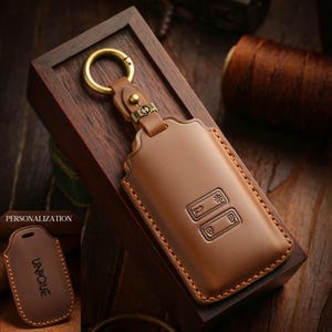 Könnte beinhalten: Eine braune Leder-Schlüsselanhänger-Hülle mit einem goldfarbenen Ring und Verschluss. Die Hülle hat geprägte Details und sichtbare Nähte. Eine kleinere Version der Hülle ist mit dem Aufdruck "UNIQUE" abgebildet. Die Schlüsselanhänger-Hülle wird in einer Holzkiste präsentiert.