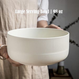 Peut inclure: Un grand bol de service avec une finition blanc cassé mate et une texture nervurée verticale sur l'extérieur. Le bol présente le texte Large Serving Bowl | 98 oz imprimé en blanc au-dessus du bord, indiquant une capacité de 98 onces.