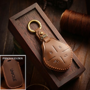 May include: A brown leather key fob cover with a gold-colored ring and clasp. The cover has stitched edges and embossed symbols. A smaller image shows a similar cover with the word "UNIQUE".
