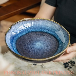 May include: A ceramic bowl with a gradient blue glaze, shifting from a deep indigo centre to lighter shades at the rim. The bowl has an irregular shape and a golden-brown edge. The text "PERSONALIZED LASER PRINTING" is at the bottom.