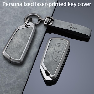 Könnte beinhalten: Ein graues, personalisiertes laserbedrucktes Schlüsselcover-Set mit einem Metallring und einem passenden Band mit dem Text Aurora. Die Cover haben eine strukturierte graue Oberfläche mit Metallrahmen und zeigen geprägte Tasten-Symbole.