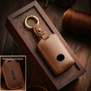 May include: A brown leather key fob case with a gold-colored ring and clasp. The case has a stitched border and a circular cutout. The word "UNIQUE" is visible on a smaller version of the case, with the word "PERSONALIZATION" above it.