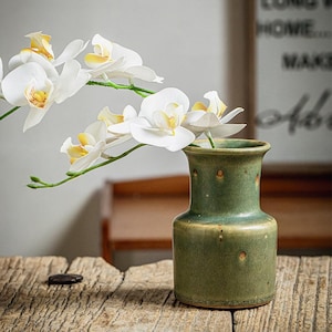 Puede incluir: Un jarrón de cerámica verde con borde acampanado y acabado de esmalte moteado, que contiene un tallo de flores de orquídea blancas con centros amarillos. El jarrón tiene una base cilíndrica y un cuello estrecho, con una textura suave y terrosa.