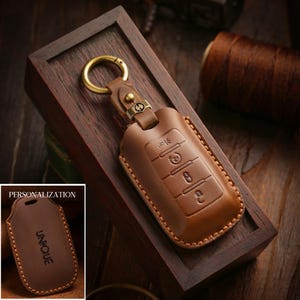 May include: A brown leather key fob case with a gold-colored keyring and clasp. The case has embossed key symbols and a stitched border. The image includes a smaller inset showing a similar case with the word "UNIQUE".