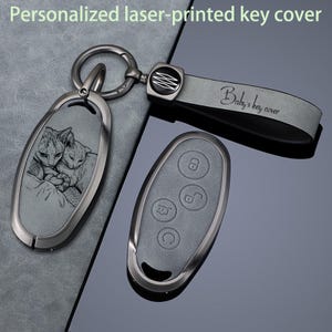Puede incluir: Una funda de llave gris con una imagen grabada con láser de dos gatos acurrucados. El conjunto incluye un anillo de metal y una correa gris con el texto "Baby's key cover" y un emblema. Se muestra un mando de llave gris con cuatro botones.