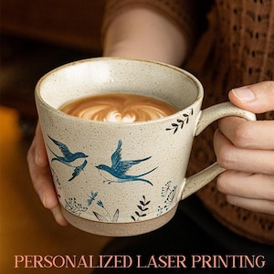 May include: A beige ceramic mug with a brown base and handle, filled with coffee. The mug is decorated with blue bird illustrations and floral accents. The text "PERSONALIZED LASER PRINTING" is at the bottom.