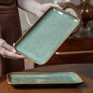Pode incluir: Dois tabuleiros de servir retangulares em cerâmica com um esmalte salpicado em verde sálvia e azul-esverdeado, com uma borda castanho-escura. A superfície tem um aspeto texturado e manchado com acabamento brilhante e bordas arredondadas.