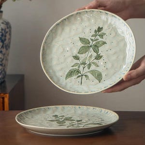 Può includere: Un piatto rotondo in ceramica con una superficie testurizzata color crema e azzurro, caratterizzato da un'illustrazione botanica verde di un fusto di pianta con foglie. Il piatto presenta un bordo leggermente irregolare e ondulato con accenti blu.