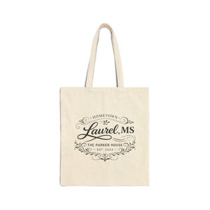 May include: Cream-colored tote bag with black text and design. The design includes the words "Hometown Laurel, MS" in a decorative font, with "The Parker House" and "Est. 2024" below. The bag has two handles.