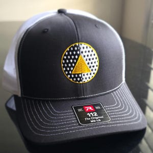 May include: A charcoal gray and white baseball cap featuring a circular patch on the front. The patch displays a black and white dotted pattern with a solid yellow triangle in the center. A black sticker on the brim reads 112 The Original MD-LG.