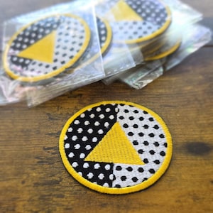 May include: Embroidered patch with a yellow triangle and black and white polka dot pattern. The patch has a yellow border and is circular in shape. Several patches are visible, some in clear plastic packaging.
