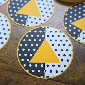 May include: Circular stickers featuring a split design with a black background with white polka dots on the left and a white background with black polka dots on the right. A solid yellow triangle is centered over the vertical dividing line.
