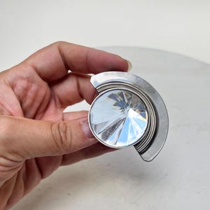 Könnte beinhalten: Ein silberfarbener Möbelknopf aus Metall mit einer halbkreisförmigen Basis und einem zentralen, runden, facettierten Glaseinsatz. Das Glaselement weist ein strahlenförmiges Muster mit reflektierenden Oberflächen auf, die das Licht einfangen.
