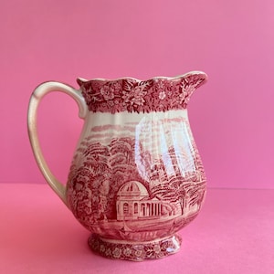 Könnte beinhalten: Vintage Keramikkrug mit rot-weißem Toile-de-Jouy-Design. Der Krug zeigt eine detaillierte Landschaftsszene mit Bäumen und einem Gebäude. Der Rand und der Boden haben florale Muster. Der Griff ist gebogen und der Krug hat einen gewellten Rand.