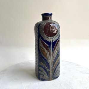 May include: A rectangular ceramic vase with a narrow neck, featuring a deep blue glaze and a prominent circular brown floral motif on the front. The sides are decorated with a textured, repeating pattern, while the base displays stylized leaf designs in shades of brown, gray, and blue.