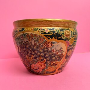 Può includere: Una ciotola decorativa in ceramica con bordo dorato e intricati disegni floreali e di pavoni. La ciotola presenta uno sfondo blu scuro con fiori colorati e una fascia dorata a motivi sulla parte superiore.