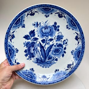 Könnte beinhalten: Ein runder Keramikteller mit weißem Untergrund und komplexen blauen Blumenmustern. In der Mitte befindet sich ein großer Blumenstrauß, während der Rand mit einer sich wiederholenden geometrischen Bordüre und floralen Motiven verziert ist.
