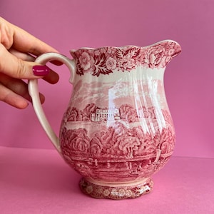Könnte beinhalten: Vintage Keramikkrug mit rot-weißem Transferdruck. Der Krug zeigt eine Landschaftsszene und einen floralen Rand. Er hat einen gebogenen Griff und einen gewellten Rand. Der Sockel hat ein dekoratives Muster.