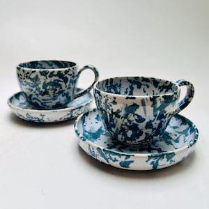 Könnte beinhalten: Zwei Keramik-Teetassen mit passenden Untertassen, die eine weiße Basis mit einem dunkelblauen, gesprenkelten Marmormuster aufweisen. Jede Tasse hat eine abgerundete Schalenform mit einem geschwungenen Henkel und jede Untertasse einen Rand.