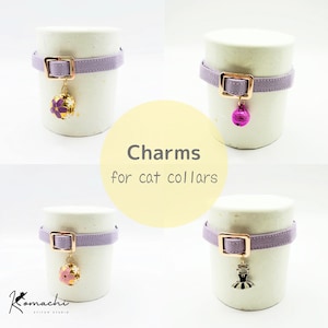 Peut inclure: Quatre colliers pour chat violet clair avec boucles en or rose, chacun doté d'une breloque unique : une sphère florale dorée, une clochette rose vif, une sphère fleur de cerisier dorée et un tutu de ballet argenté. Texte : Charms for cat collars.
