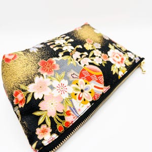 May include: A black zippered pouch featuring a vibrant floral pattern with pink, white, and red blossoms, accented by gold-colored metallic details and stylized circular motifs. The rectangular accessory has a gold-toned zipper pull.