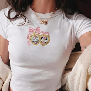 May include: White t-shirt featuring a pink bow and two heart-shaped lockets embroidered with cartoon characters. The top has short sleeves and a scoop neck. A gold necklace with a heart pendant is also visible.