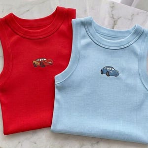 May include: Two ribbed tank tops, one red and one light blue, each with a small embroidered car design. The tops are sleeveless and have a round neckline. The red top has a yellow and red car, and the blue top has a blue car.