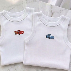 May include: Two white ribbed tank tops with embroidered cartoon car designs. One tank top features a red car, the other a blue car. Both tops have a round neckline and sleeveless design, perfect for warm weather.