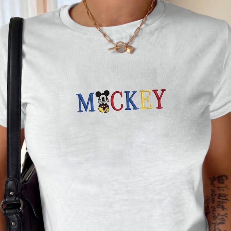 以下が含まれることがあります： 白のTシャツ。青、黄色、赤の糸で「Mickey」の文字が刺繍されています。「O」の文字はミッキーマウスのグラフィックに置き換えられています。Tシャツはクルーネックで半袖です。