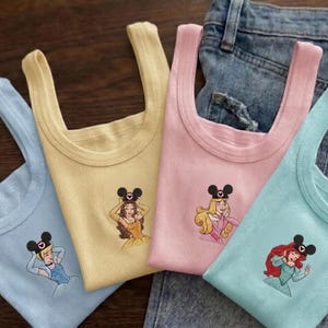 Puede incluir: Cuatro camisetas sin mangas acanaladas en colores pastel: azul claro, amarillo, rosa y turquesa. Cada camiseta presenta un bordado de una princesa de Disney con una diadema de orejas de Mickey Mouse. Las camisetas se muestran sobre un fondo de mezclilla.